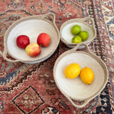 A set of 3 Respiin jute handled bowls on top of a decorated rug with fruit in each bowl