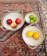 A set of 3 Respiin jute handled bowls on top of a decorated rug with fruit in each bowl