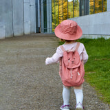 A toddler wearing the Roommate kids canvas backpack in pink with a bunny design, and a matching pink bucket hat.