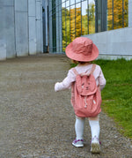 A toddler wearing the Roommate kids canvas backpack in pink with a bunny design, and a matching pink bucket hat.