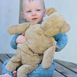 A young child holding the large Senger beige bear, the teddy is facing the child and it's back is facing the camera.