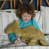 A child sitting up in bed with a large Senger cuddly camel soft toy placed on their lap. The child is looking down at the camel with a smile on his face.