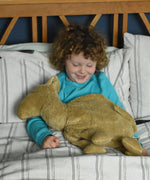 A child sitting up in bed with a large Senger cuddly camel soft toy placed on their lap. The child is looking down at the camel with a smile on his face.