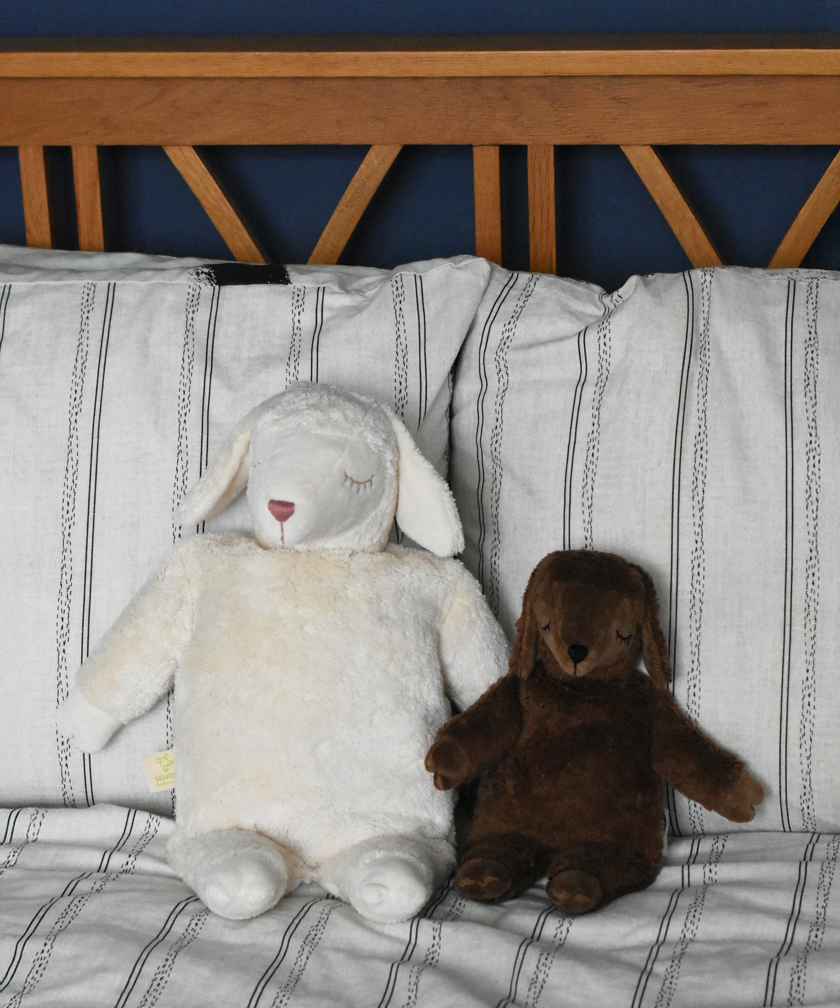 A Senger large white sheep and small brown sheep soft toy in seated position next to each other, leaning against pillows and a wooden headboard. These sheep are part of a wide range of soft toys made from organic and natural materials stocked at Babipur.