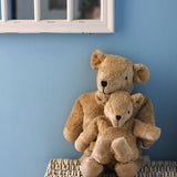 A small Senger cuddly beige bear sat on the lap of a large bear. They are sitting on top of a white wicker laundry basket in front of a blue wall