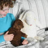 A Senger small brown cuddly sheep soft toy in placed on a child's lap in bed. The child is holding one of the sheep's hands. A large white cuddly sheep can be seen next to the child. These sheep are part of a wide range of soft toys made from organic and natural materials stocked at Babipur.