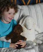 A child holding the Senger small brown cuddly sheep soft toy on their  lap in bed. A large white cuddly sheep can be seen next to the child. These sheep are part of a wide range of soft toys made from organic and natural materials stocked at Babipur.