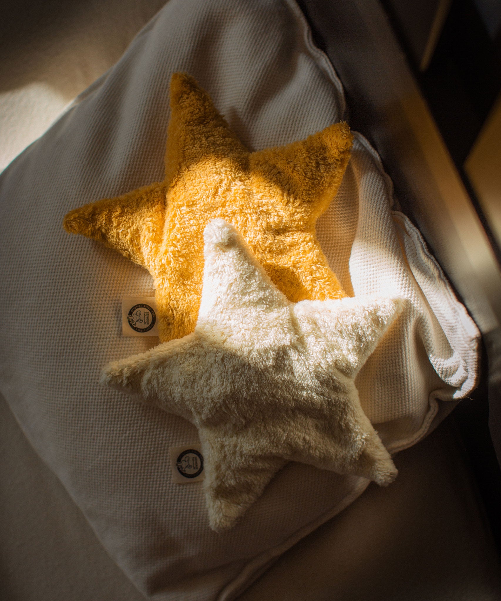 Two Senger star shaped warming cushions placed on a bed. One is a light yellow, off white colour and the other is a bright yellow colour.