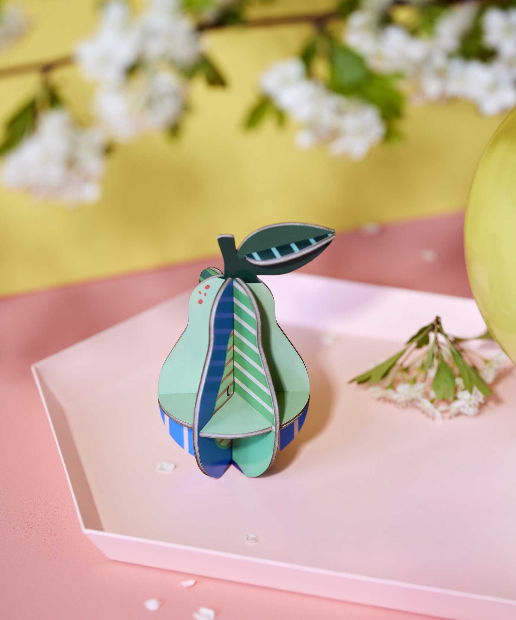 The Studio Roof 3D Pop Out Card Pear placed on a pink tray next to a yellow vase. This 3D pop out card DIY kit is part of Studio Roof's fruits collection which is available at Babipur.