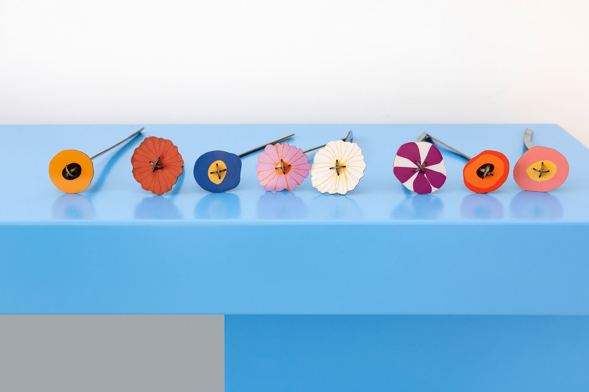 The 8 flowers from the Studio Roof Cardboard Bouquet in a  Klimt's Garden design placed on a blue surface all lined up. A DIY Cardboard craft kit that makes up 8 3D flowers. This 3D pop out DIY kit is part of Studio Roof's artist flowers collection which is available at Babipur.