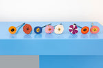 The 8 flowers from the Studio Roof Cardboard Bouquet in a  Klimt's Garden design placed on a blue surface all lined up. A DIY Cardboard craft kit that makes up 8 3D flowers. This 3D pop out DIY kit is part of Studio Roof's artist flowers collection which is available at Babipur.