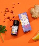 Think Positive pure essential oil blend in a blue glass bottle next to a decorative purple cardboard box, with peppercorns, ginger, lemon and geranium on an orange background.