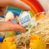 Close up of a hand holding a Tonys chocolonely fairtrade mini easter egg in front of some wooden chick toys