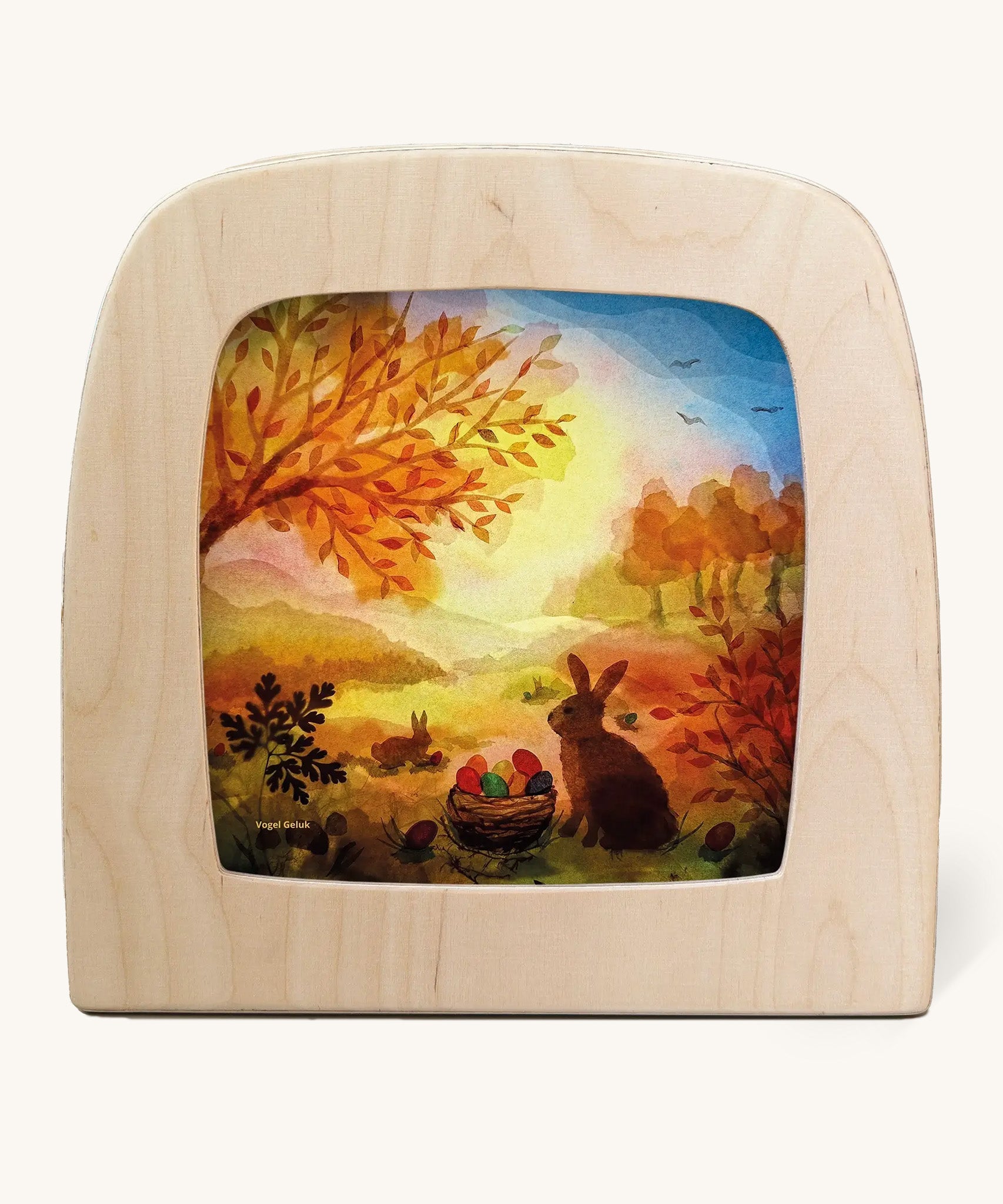 A Toverlux silhouette inside a wooden  lamp. This silhouette is Spring inspired with a rabbit standing next to a basket full of coloured eggs under a tree This layered paper style silhouette is by artist Vogel Geluk and available at Babipur.
