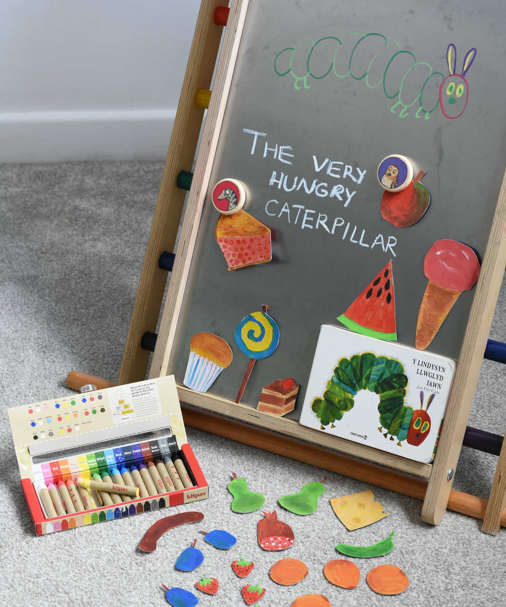 A Triclimb Biri placed on a rainbow Triclimb. A pack of Kitpas crayons have been placed on the floor in front of it. The board has been decorated with hand drawn food illustrations placed under Bajo gruffalo magnets and a caterpillar featured in the very hungry caterpillar book.