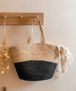 The Turtle Bags hand woven and fair trade jute basket bag with handles, and a black colour block design, hanging on a peg