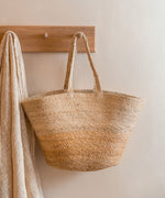 The Turtle Bags hand woven and fair trade jute basket bag with handles, and a natural colour block design, hanging on a peg