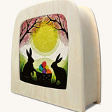 A Toverlux silhouette inside a lamp with 2 bunny rabbits standing next to a basket full of coloured eggs under a blossom tree. This layered paper style silhouette is by artist Vogel Geluk and available at Babipur.