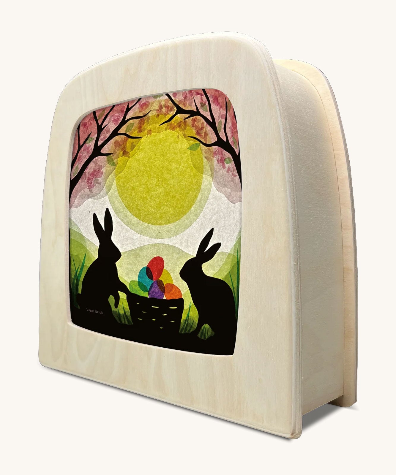 A Toverlux silhouette inside a lamp with 2 bunny rabbits standing next to a basket full of coloured eggs under a blossom tree. This layered paper style silhouette is by artist Vogel Geluk and available at Babipur.