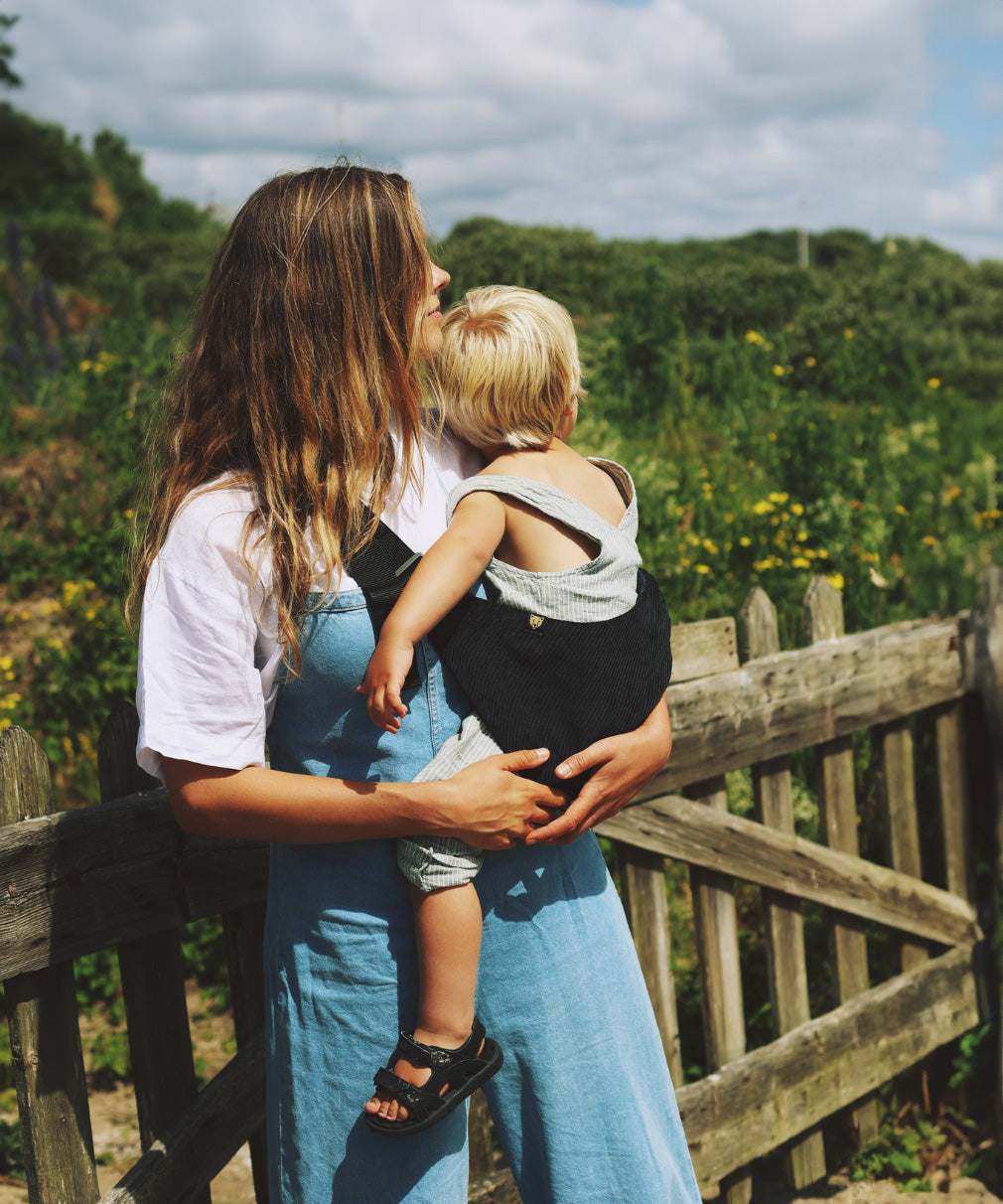 A woman carrying her toddler in the Wildride black corduroy toddler carrier, with lush grass in the background.