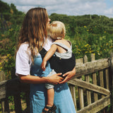 A woman carrying her toddler in the Wildride black corduroy toddler carrier, with lush grass in the background.