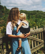 A woman carrying her toddler in the Wildride black corduroy toddler carrier, with lush grass in the background.