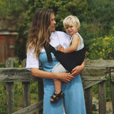 A woman carrying her toddler in the Wildride black corduroy toddler carrier, with lush grass in the background.
