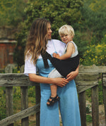 A woman carrying her toddler in the Wildride black corduroy toddler carrier, with lush grass in the background.