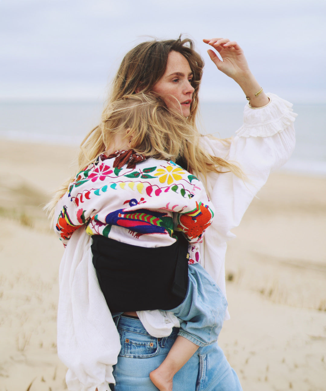 A woman on a breezy beach, carrying her toddler in the Wildride ergonomic Black Linen toddler carrier sling.