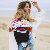 A woman on a breezy beach, carrying her toddler in the Wildride ergonomic Black Linen toddler carrier sling.