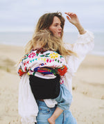 A woman on a breezy beach, carrying her toddler in the Wildride ergonomic Black Linen toddler carrier sling.
