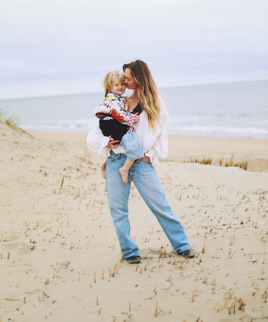 A woman on a sandy beach, carrying her toddler with the support of the Wildride black linen toddler carrier sling.
