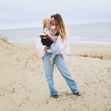 A woman on a sandy beach, carrying her toddler with the support of the Wildride black linen toddler carrier sling.