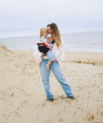 A woman on a sandy beach, carrying her toddler with the support of the Wildride black linen toddler carrier sling.