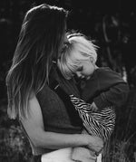 A mother carrying a toddler closely in the black zebra stripe toddler Wildride carrier showing the strap and seat detail