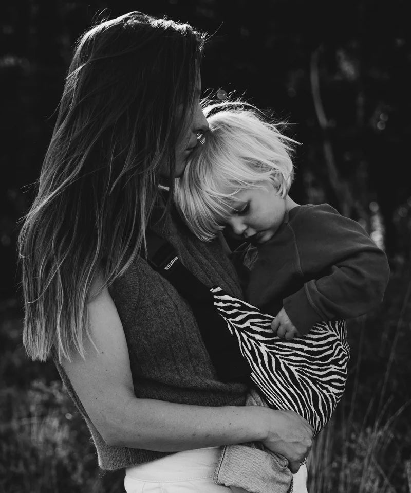 A mother carrying a toddler closely in the black zebra stripe toddler Wildride carrier showing the strap and seat detail