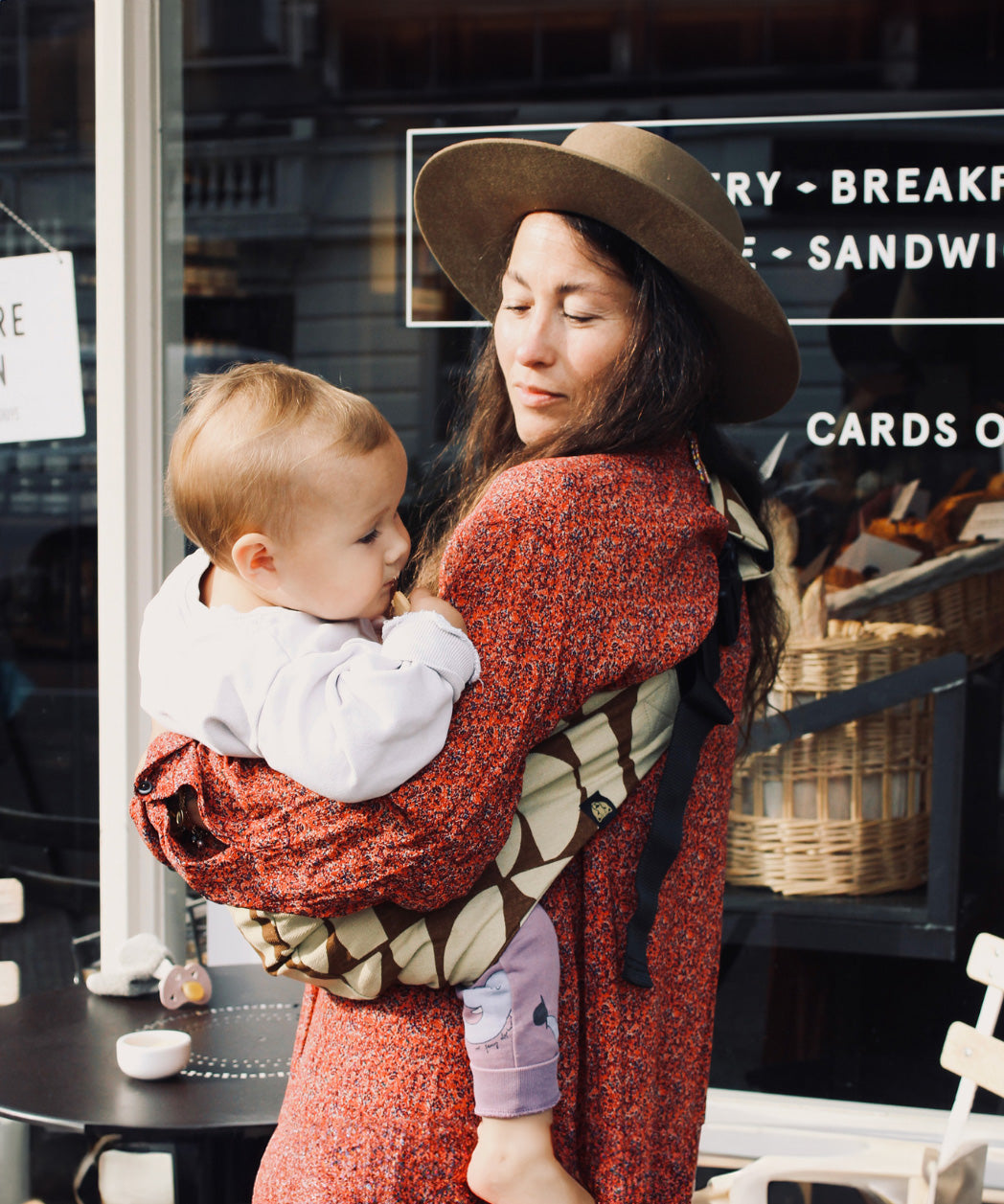 A woman outside a café, carrying her baby in the stylish Wildride hip sling carrier, in a Boho design