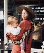 A woman outside a café, carrying her baby in the stylish Wildride hip sling carrier, in a Boho design
