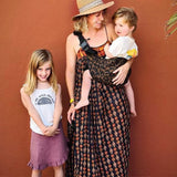 A mother carrying a toddler in the brown leopard Wildride toddler carrier with a child standing to them showing the size of the seat and strap buckle detail.