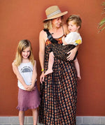 A mother carrying a toddler in the brown leopard Wildride toddler carrier with a child standing to them showing the size of the seat and strap buckle detail.