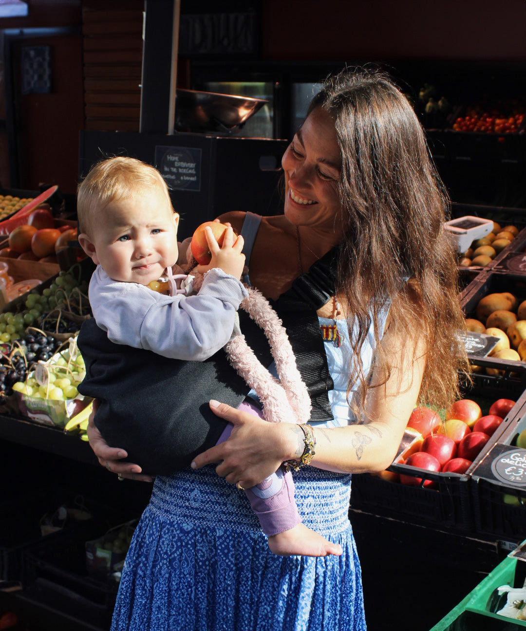 A woman in a fruit market, holding her baby with support from the Wildride toddler hip sling carrier, in charcoal grey.