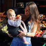 A woman in a fruit market, holding her baby with support from the Wildride toddler hip sling carrier, in charcoal grey.