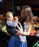 A woman in a fruit market, holding her baby with support from the Wildride toddler hip sling carrier, in charcoal grey.