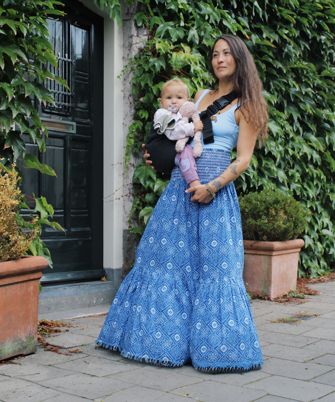 A woman in a long skirt holding her baby with support from the Wildride toddler hip sling carrier, in charcoal grey.