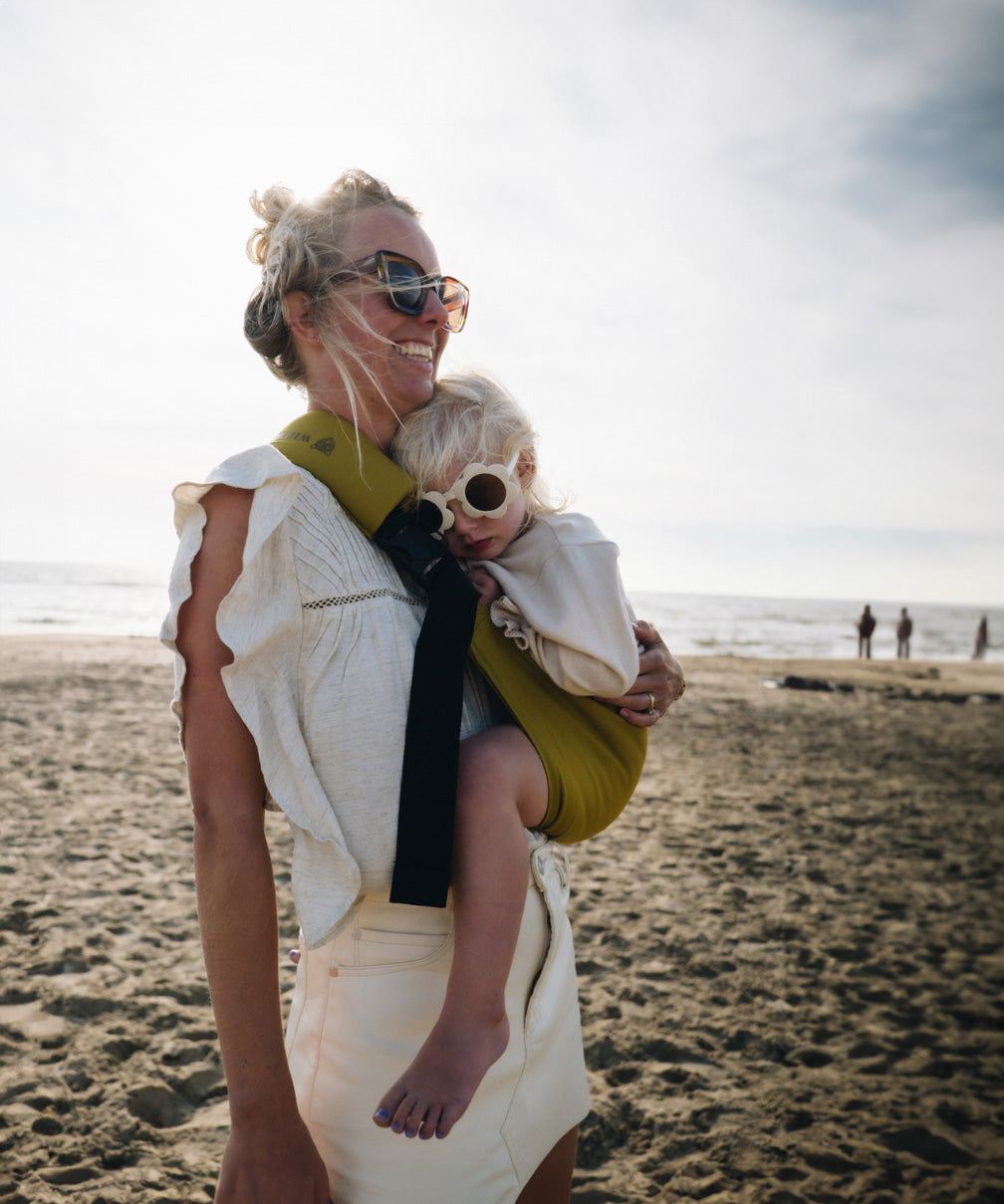 A woman in sunglasses on the beach, carrying her toddler (also in flower shaped sunglasses) in the Wildride toddler hip sling carrier in desert yellow