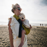 A woman in sunglasses on the beach, carrying her toddler (also in flower shaped sunglasses) in the Wildride toddler hip sling carrier in desert yellow