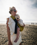 A woman in sunglasses on the beach, carrying her toddler (also in flower shaped sunglasses) in the Wildride toddler hip sling carrier in desert yellow
