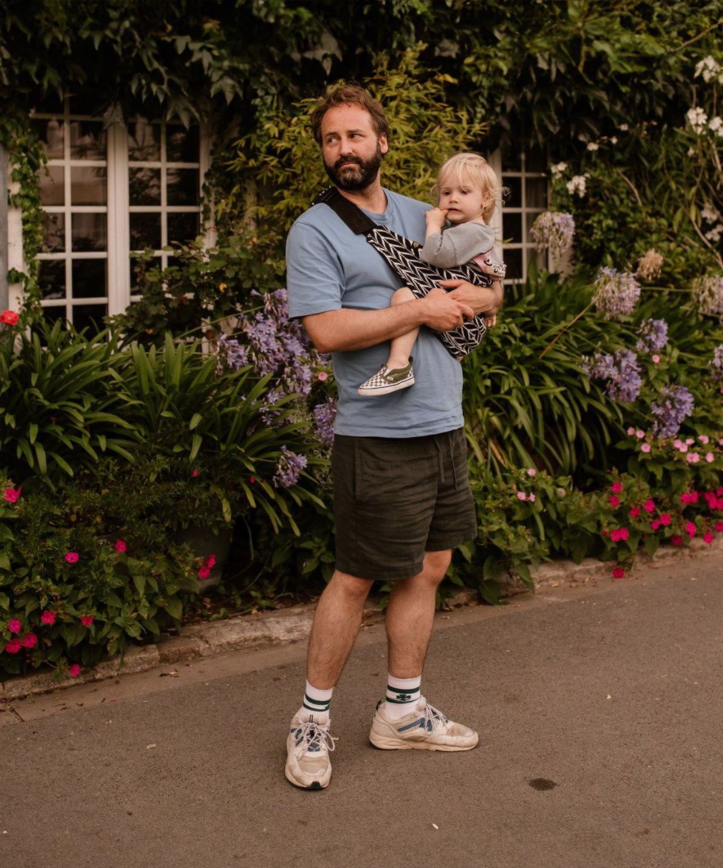 A man carrying his toddler in the special edition 'Feathers' print Wildride hip sling carrier, in front of plants