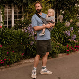 A man carrying his toddler in the special edition 'Feathers' print Wildride hip sling carrier, in front of plants