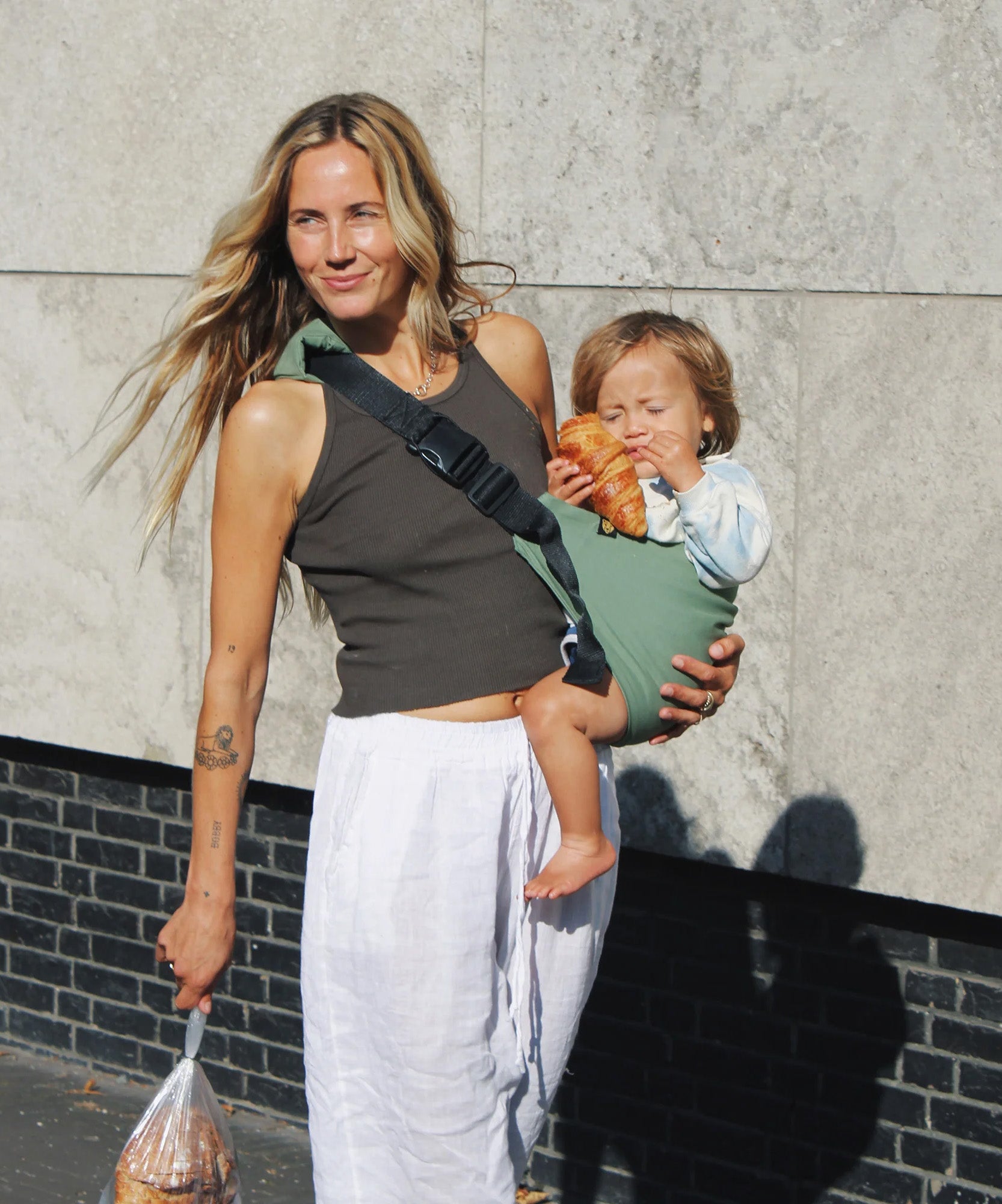 A woman carrying her toddler in the Wildride carrier, in a green fern colour